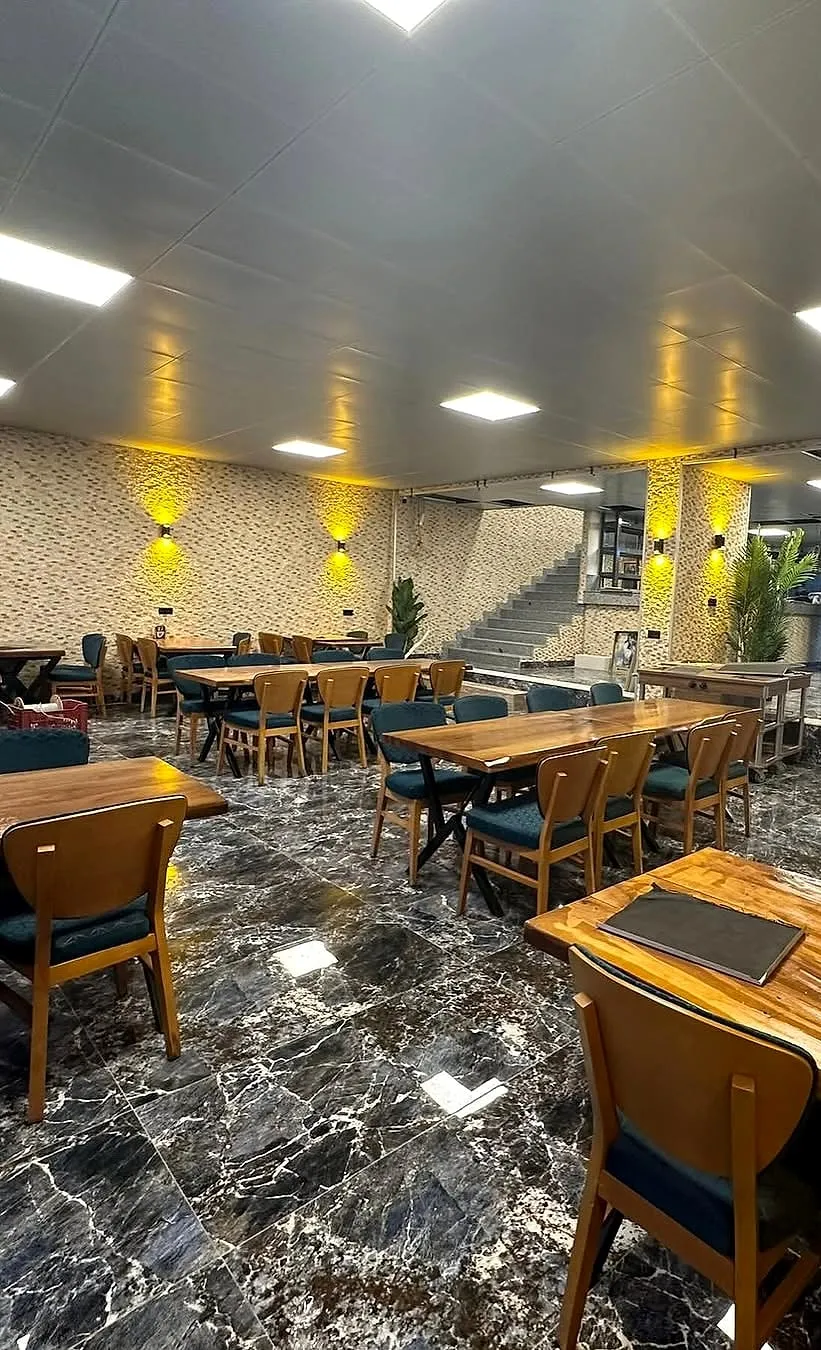 Restoran salonu: ahşap masalar ve petrol mavisi döşemeli sandalyeler, aplikli taş görünümlü duvar kaplaması, clip-in tavanda kare LED paneller ve koyu mermer görünümlü zemin