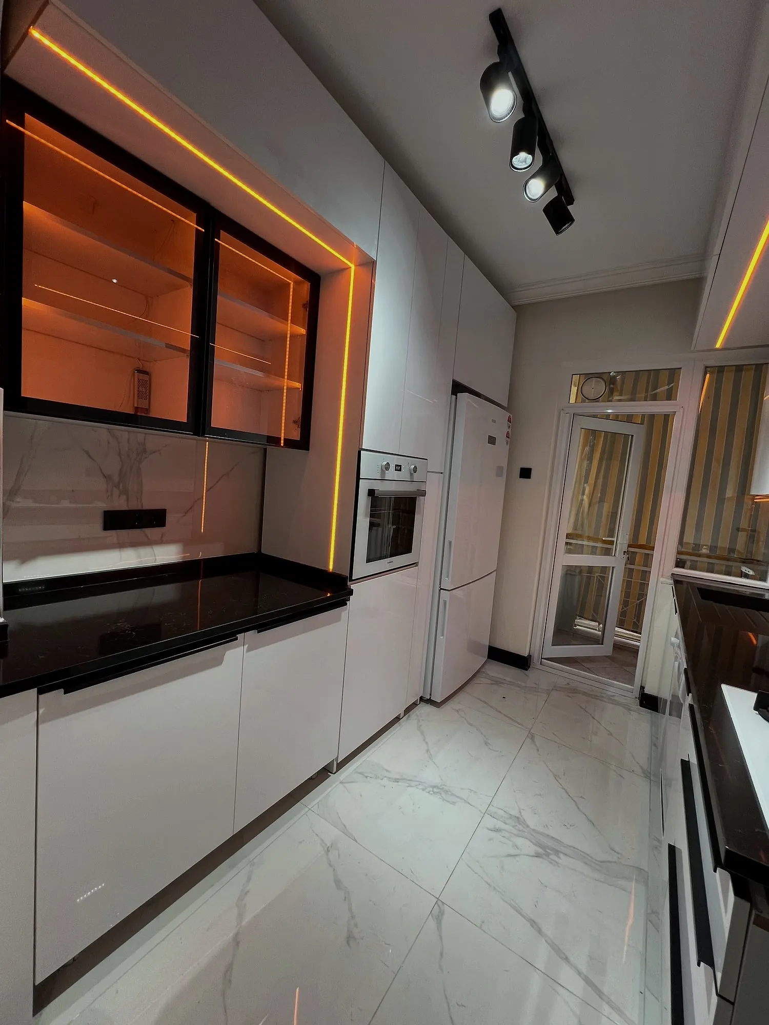 Balkon kapısına uzanan koridor mutfak; solda kehribar ışıklı cam dolaplar, sağda beyaz fırın ve buzdolabı